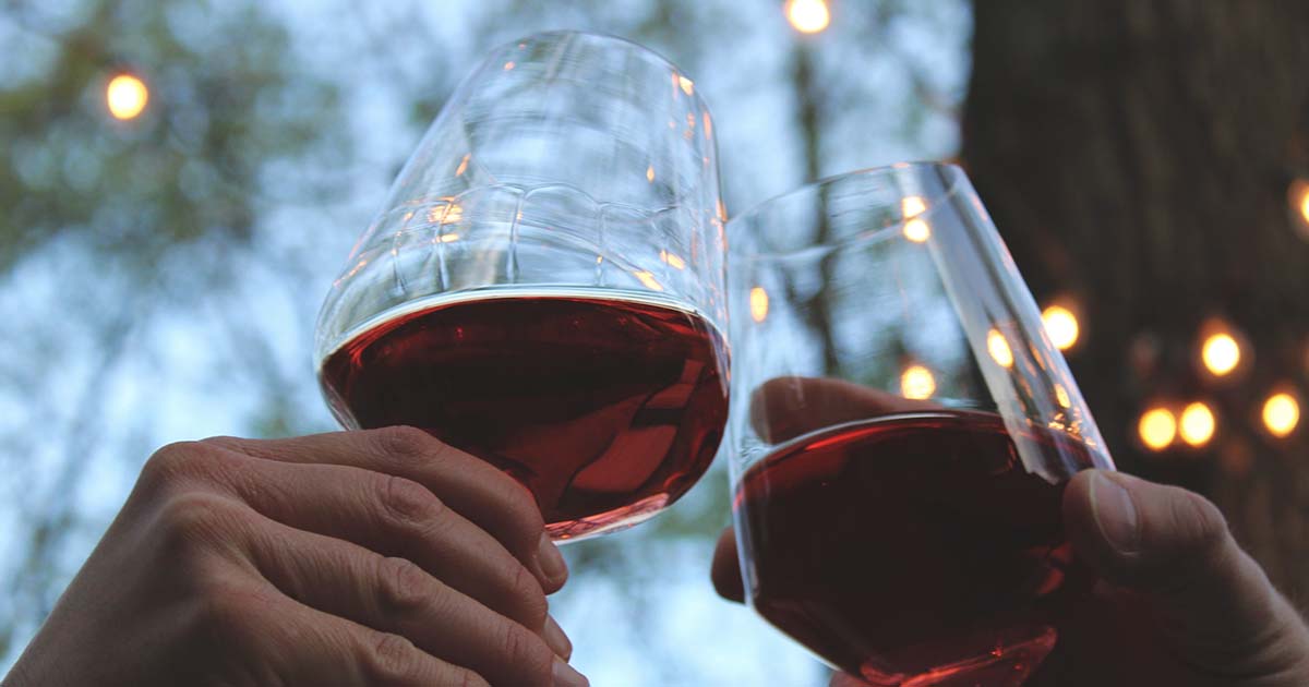 Two glasses of red wine being held by two people outside with lights